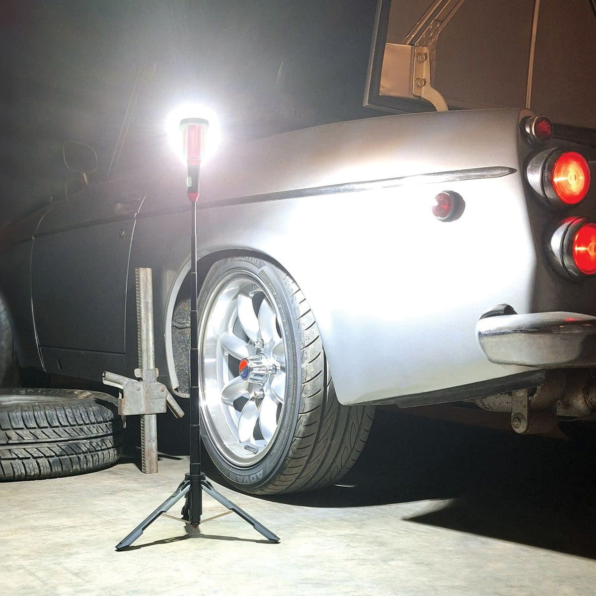 Car with a spare tire and jack nearby in a garage setting, lit up by a FLi-MORPHE Telescoping Light by STKR